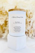 Load image into Gallery viewer, A matte white glass jar candle with a natural bamboo lid, labeled 'Rose Petals' for a rose-scented candle. The candle is displayed on a white stand against a backdrop of dry grass, with a wooden wick visible.
